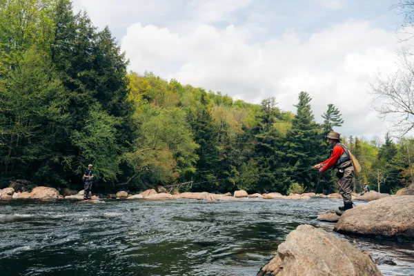 Two fly fishermen in waders and vests stand on large river rocks casting their lines into a wide, flowing stream.