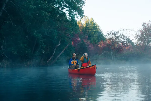 Man paddles at back of red canoe on foggy river while woman in front drinks from green thermos, both wearing life vests