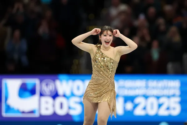 Woman in a gold dress smiles and raises arm in celebration while performing on ice at figure skating competition