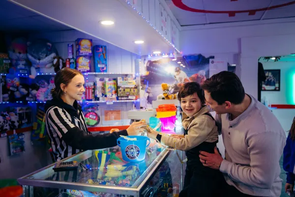 Woman in black and white striped ref-style top hands something across counter to small boy held by father at arcade