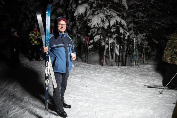 Person standing outdoors at night in cold weather clothing holding cross-country skis in one hand and a cup in the other