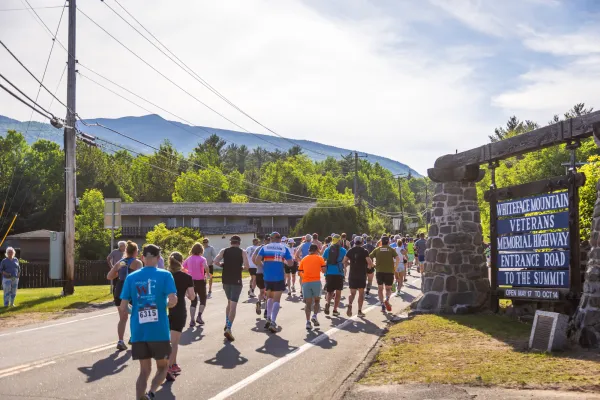A large group of runners head down a road, passing a stone sign for the Whiteface Mountain Veterans Memorial Highway.