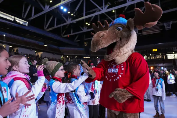 A group of children in colorful winter jackets high-five a person in a cheerful moose mascot costume at an indoor event, conveying excitement and joy.