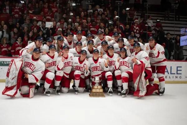 Women's hockey team in uniform and ECAC championship hats gathered for large group photo on ice with trophy