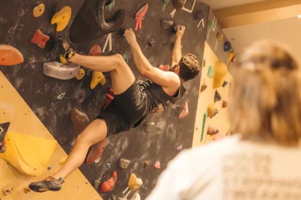 Bouldering climber positioned sideways while ascending a route on an indoor wall while another person looks on