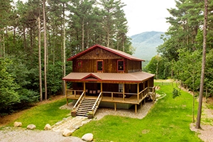 A two story cabin with red trim, a wrap around covered porch, lush lan and a perfect view of a mountain through the tall trees