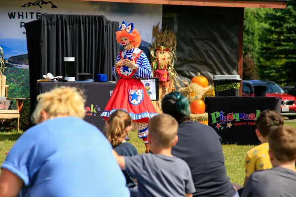 Woman dressed as a clown stands at front of a crowd of children sitting on grass talking to them