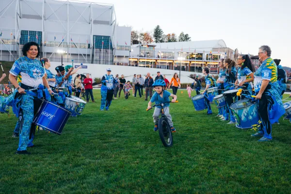 Child rides bike through aisle created by drum line on grass of Olympic Speed Skating Oval