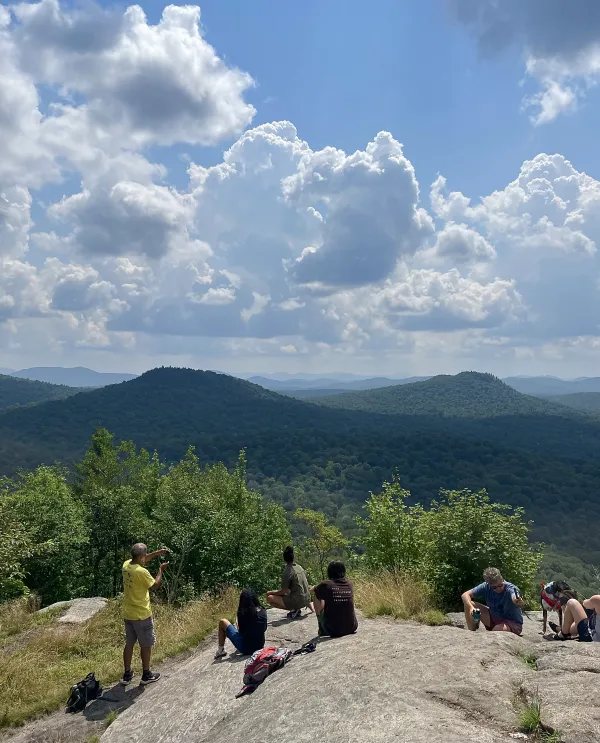 Hikers sit and stand on rocky summit of Goodman Mountain looking at and photographing the view.