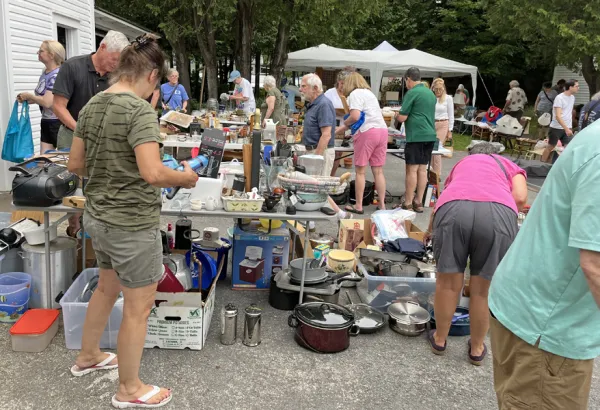 People browse outdoor bazaar sale with items on tables, on the ground, and under pop-up tents