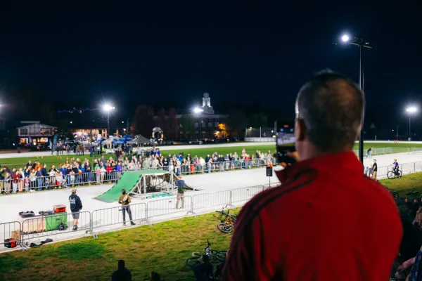 Man takes video of BMX stunt show rider performing a flip off a ramp at the Olympic Oval under the lights at night