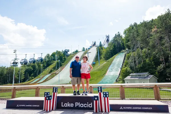 A man and woman pose on a Lake Placid podium in front of Olympic ski jumps on a sunny summer day.