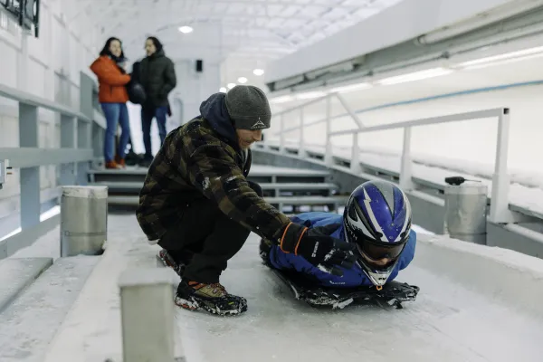 Track worker provides instruction to person in helmet laying on skeleton sled on bobsled, luge, and skeleton track