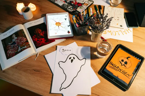Stock image of table with art supplies, paper with drawing of a ghost, photobooks lying open, and a tablet with a digital halloween drawing