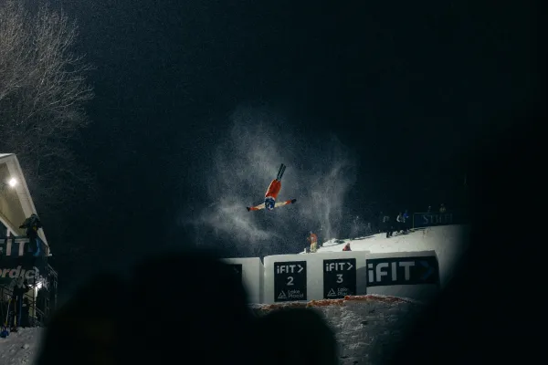 Freestyle aerialist skier performs a flip after skiing off a jump at a nighttime competition under bright lights while a crowd looks on