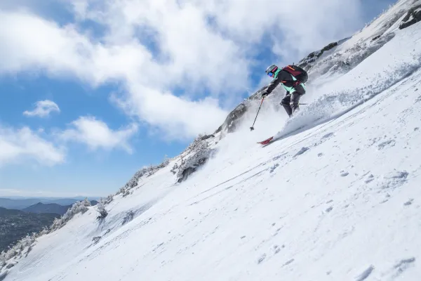Skier in a red jacket descends a steep, snowy mountain slope under a bright blue sky. The scene conveys an exhilarating sense of adventure and speed.