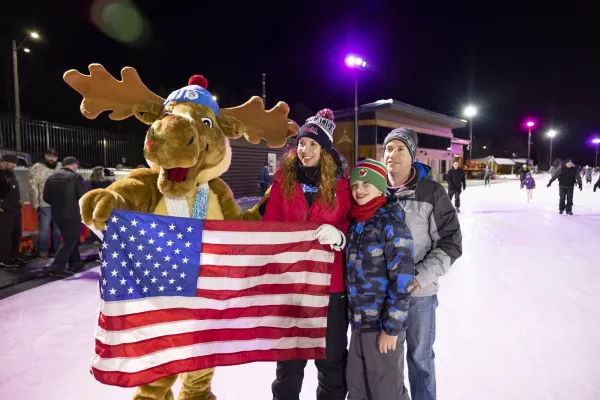Family stands with moose mascot holding American flag on Olympic Speed Skating Oval at night taking group picture