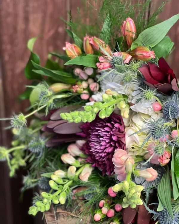 A bouquet of pink snap dragons, blue sea holly, and and burgundy daisies surrounded with assorted foliage accents lays on a table.