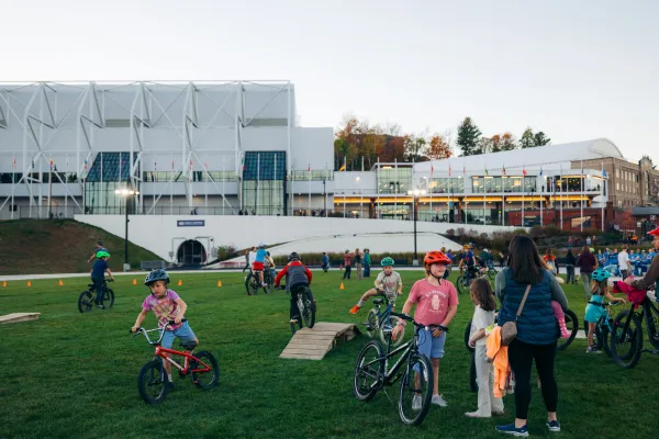 Kids ride on small mountain bike features on grassy area of Olympic Oval in Lake Placid with Olympic Center in background