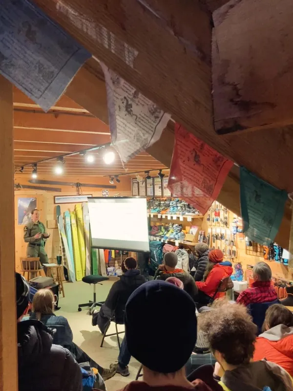 Forest Ranger gives a presentation next to a white screen to a room of sitting people with prayer flags hanging from the ceiling