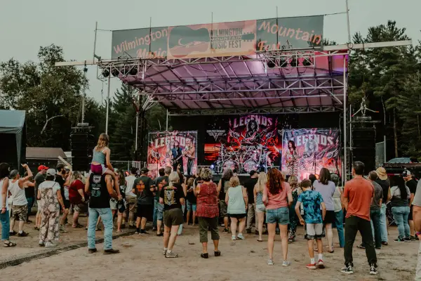 Crowd of people stand on dirt lawn in front of temporary music stage  in summer