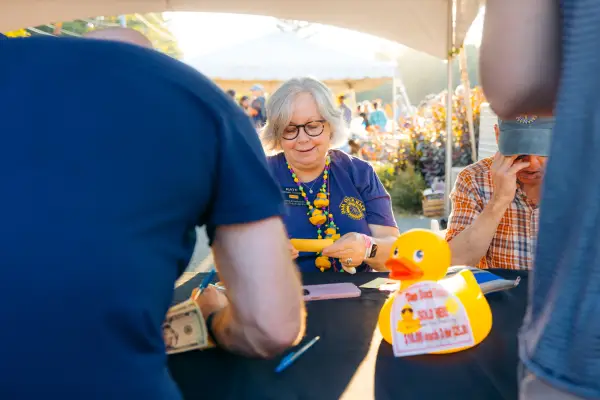 Woman with grey hair and glasses wearing blue Rotary shirt and duck accessories is seen selling something 