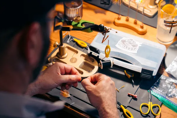 A person’s hands are shown as they meticulously tie a fly on a green and gold rotary vise at a wooden workbench.