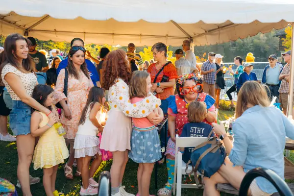 Children and parents wait in line looking on as seated clown paints the face of a child 