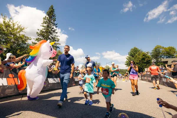 Group of kids and parents, some filming, walk in Ironkids race course past inflatable unicorn mascot