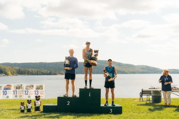Three male runners stand on a three-tiered winner's podium by a lake, each holding a carved bear trophy.