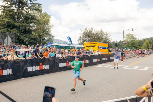 Child in teal shirt, blue shorts, and visor runs on Ironkids course while spectators cheer from behind Ironman fencing