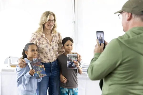 Two kids stand on either side of woman, each of them holding up a book while a man takes their photo with a cell phone