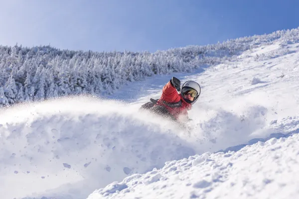 Snowboarding in red jacket, helmet, and goggles sprays snow as they descend a steep, powdery, ungroomed slope