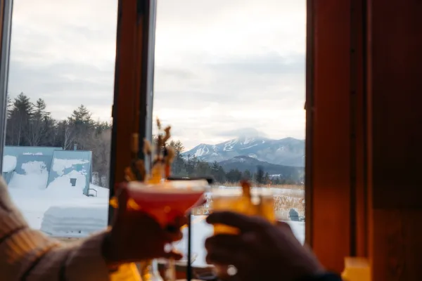 Two hands toast with cocktails in front of a window overlooking a snowy mountain landscape at dusk.