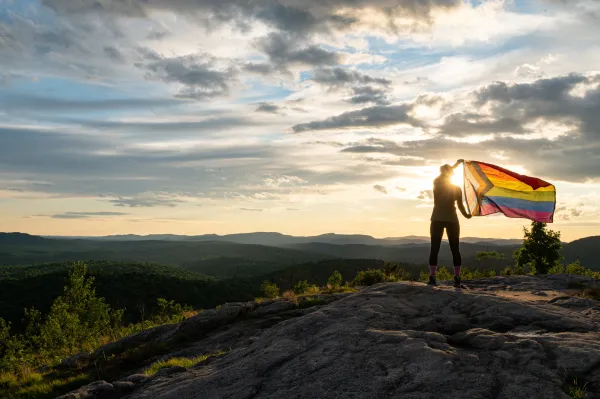 A person stands on a rocky hilltop, holding a brightly colored pride flag against a scenic sunset. The sky is dramatic with scattered clouds.
