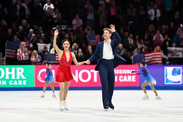 Pair of figure skaters, a woman in a red dress and a man in a blue suit, wave to crowds from on the ice while young girls in blue dresses skate behind