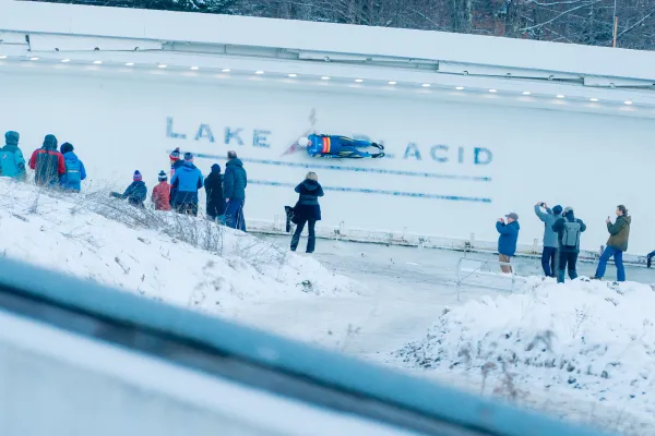 Luge athlete rides down track over where Lake Placid is printed into the ice while several people look on, some taking photos