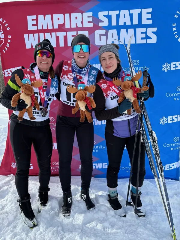 Three skiers proudly pose in front of the "Empire State Winter Games" banner, holding moose plushies and medals, exuding joy and accomplishment in snowy attire.