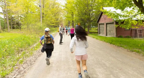 A group of people hiking on a gravel path through a wooded area, with a red barn in the background.
