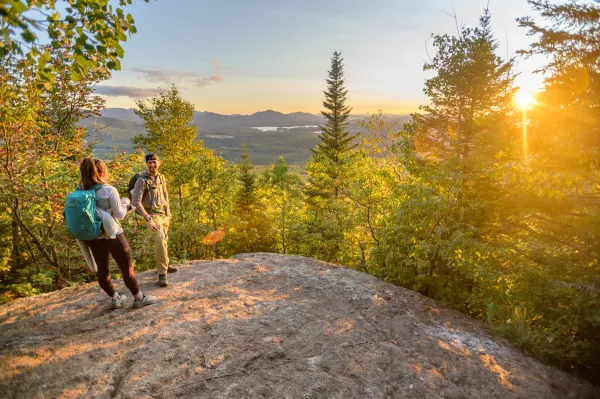 A couple talks to each other at the summit of Haystack Mountain while the sun is setting in the background