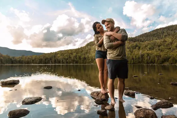 couple-embracing-at-waters-edge-with-mountains-and-blue-skies-in-the-background