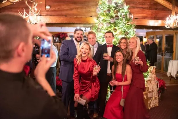 Man takes photo on a cell phone of a group of young people, women in formal dresses and men in suits, gathered together smiling in front of a decorated and lit Christmas tree