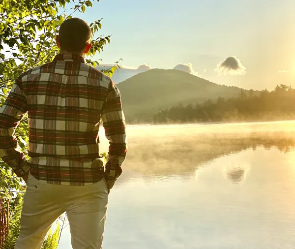 A man in a flannel looks out over a misty lake and mountains.