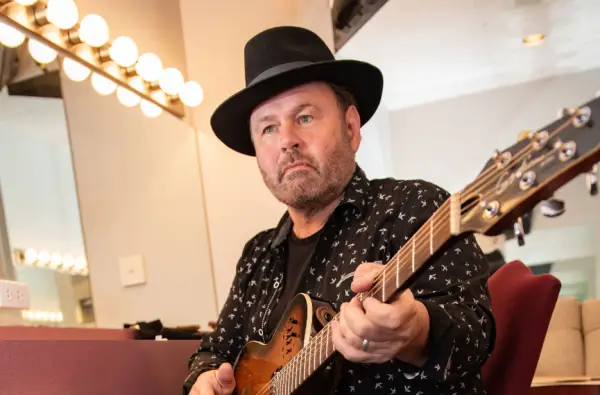 A middle-aged man with a beard wearing a black hat and a black shirt with white birds on it plays an acoustic guitar.