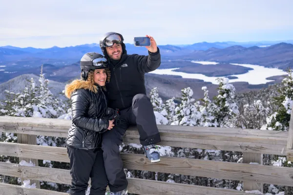 A woman and man in ski clothing and helmets take a selfie of themselves posing on a railing overlooking mountains and lakes.