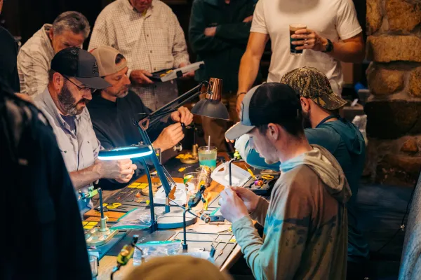 Group of people work on tying fishing flies under small lamps around a table at the Ausable River Two-Fly event