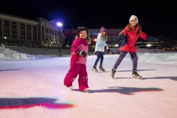 Little girl in pink snowsuit and helmet smiles at camera as she and 2 others skate by on Olympic Speed Skating Oval at night