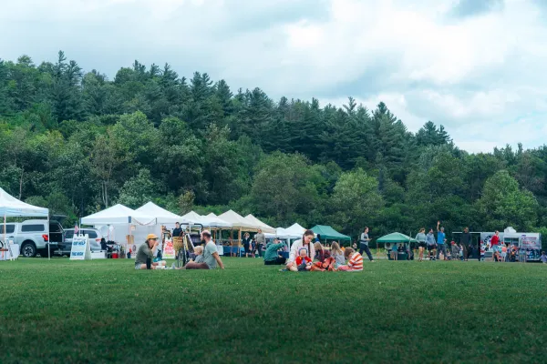 A wide shot of people enjoying a grassy field at an outdoor market with white vendor tents and a backdrop of pine trees.