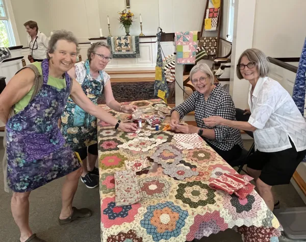 Four senior woman gathered around a table covered in a quilt and quilting materials look at camera smiling
