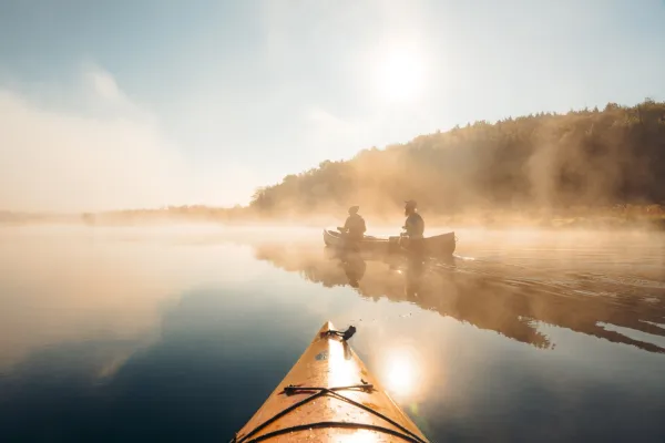 Two people paddle a red canoe through mist on mirror-like river with front of orange kayak visible in center foreground